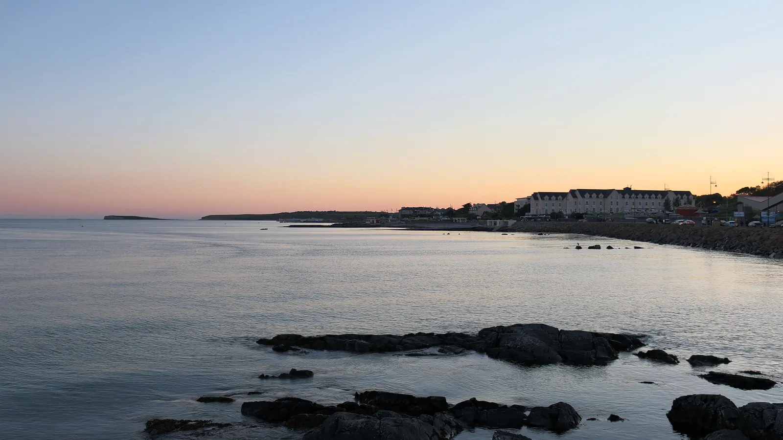 Dark rocks in the calm water of Galway Bay at dusk, with the lit buildings of the Salthill seafront along the far shore