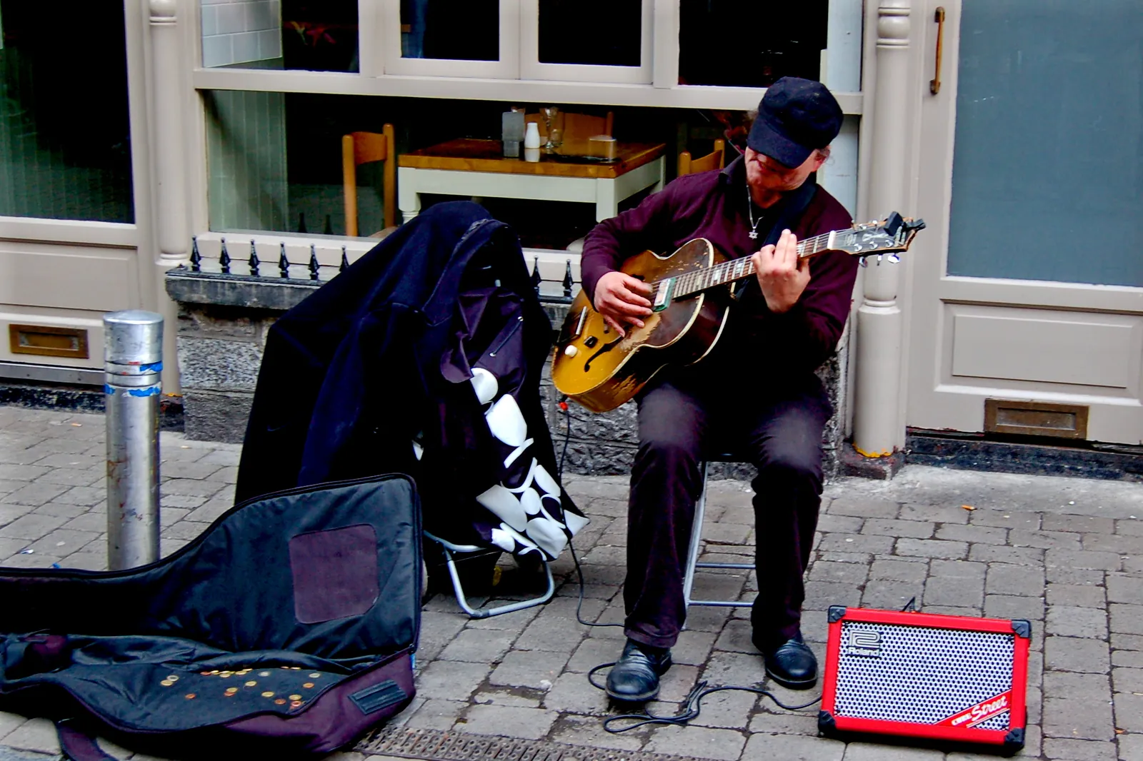 A street musician playing an acoustic guitar on a paved street in Galway, with an open guitar case and a small amplifier in front of him