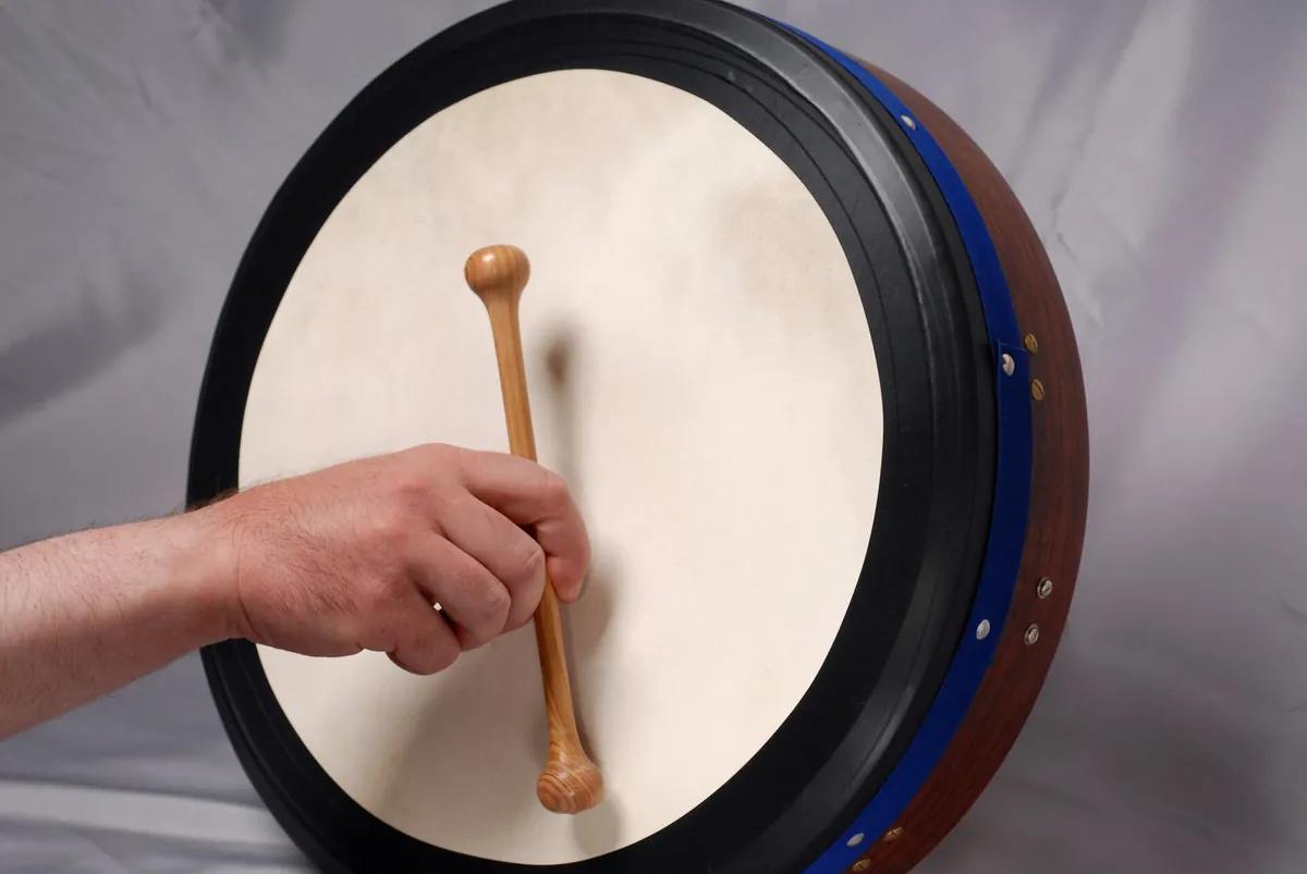 A bodhrán with a pale goatskin head and a blue rim, a hand holding a wooden beater against the skin