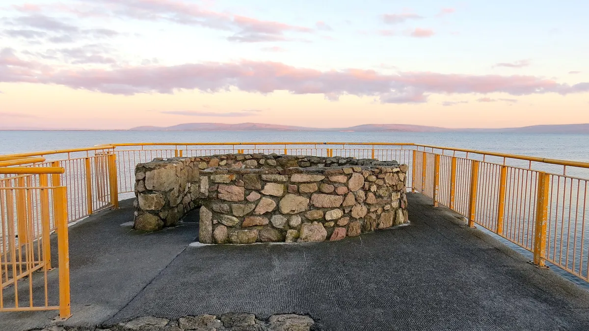 Salthill promenade at dusk, a stone-walled viewpoint behind yellow railings with Galway Bay and the hills of County Clare under a pink sky