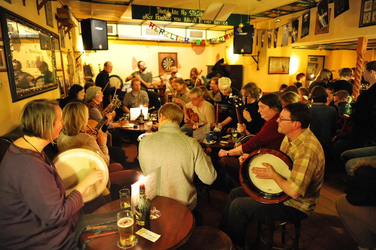 Musicians playing fiddles and guitars at a table in a pub, with glasses and a wooden counter in the background