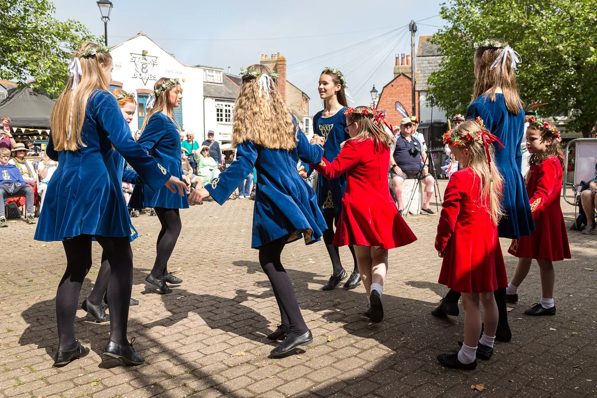 Young dancers in blue and red velvet dresses with gold Celtic knotwork and flower crowns circling hand in hand on a paved square while spectators watch
